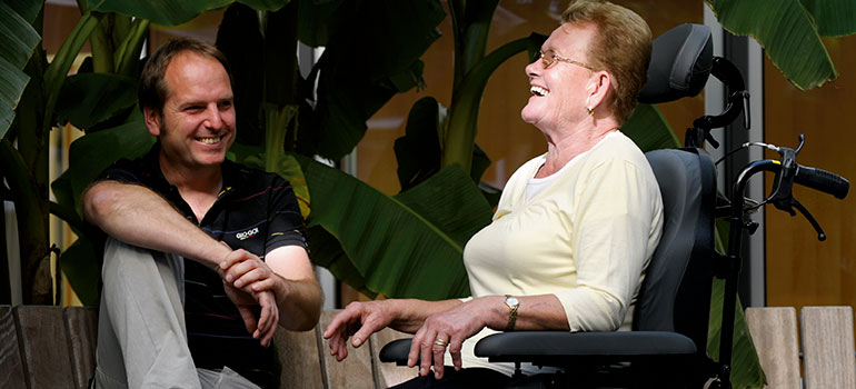 Oudere vrouw in haar rolstoel in gesprek met haar zoon.