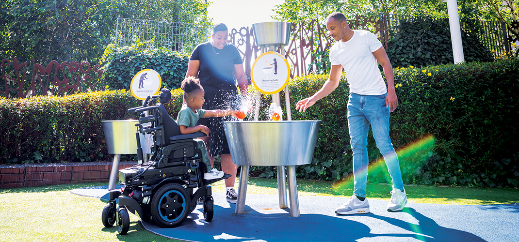 Een kind in een elektrische rolstoel die met zijn ouders aan het spelen is in een buitenspeeltuin.